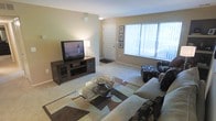 This single-floor space centers around a well-appointed living room that combines functionality with a relaxed, comfortable ambiance. The room is finished with wall-to-wall carpeting in a neutral beige tone, which complements the warm, light-colored walls throughout. A large window fitted with vertical blinds runs along one wall, allowing ample natural light to fill the space and creating a bright, airy feel.

The living room is thoughtfully arranged with a plush sofa and coordinating patterned accent chair and ottoman, all positioned around a round glass-topped coffee table. A bold geometric area rug in earth tones anchors the seating arrangement, adding visual interest to the space. Opposite the seating area, a dark wood media console houses a flat-screen television along with media storage, flanked by glass-door cabinet compartments. A table lamp atop the console provides warm ambient lighting.

A white paneled entry door is visible from the living area, framed by a trio of wall art pieces. The overall layout is open and practical, offering a welcoming environment suitable for both relaxing and entertaining.
