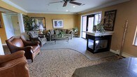 This single-floor space presents a well-appointed lounge or common area designed for comfort and sociability. The room features warm golden-yellow walls accented by white crown molding, lending the space a classic, refined character. A ceiling fan with integrated lighting serves as a functional centerpiece overhead.

The seating area is thoughtfully arranged, anchored by a patterned area rug layered over neutral carpeting. Plush upholstered sofas and armchairs, including a rich brown leather accent chair, provide ample seating options arranged around a glass-top coffee table. The furnishings are complemented by decorative indoor plants that add a natural, refreshing touch throughout.

Natural light filters into the space through a large glass door, offering a view of the exterior and brightening the interior. A dark-finished display cabinet or counter unit is positioned near the entrance, topped with floral and plant arrangements that enhance the welcoming ambiance.

Framed artwork adorns the walls, contributing to the polished, hospitality-oriented aesthetic. Overall, the space conveys a warm, comfortable environment suited for relaxation and social interaction.