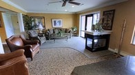 This single-floor space presents a well-appointed lounge or common area designed for comfort and sociability. The room features warm golden-yellow walls accented by white crown molding, lending the space a classic, refined character. A ceiling fan with integrated lighting serves as a functional centerpiece overhead.

The seating area is thoughtfully arranged, anchored by a patterned area rug layered over neutral carpeting. Plush upholstered sofas and armchairs, including a rich brown leather accent chair, provide ample seating options arranged around a glass-top coffee table. The furnishings are complemented by decorative indoor plants that add a natural, refreshing touch throughout.

Natural light filters into the space through a large glass door, offering a view of the exterior and brightening the interior. A dark-finished display cabinet or counter unit is positioned near the entrance, topped with floral and plant arrangements that enhance the welcoming ambiance.

Framed artwork adorns the walls, contributing to the polished, hospitality-oriented aesthetic. Overall, the space conveys a warm, comfortable environment suited for relaxation and social interaction.