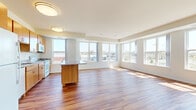 This single-floor unit is designed with an open-concept layout that seamlessly connects the kitchen and living areas, creating a spacious and airy atmosphere. Rich hardwood flooring extends throughout the main living spaces, complementing the warm-toned cabinetry found throughout the unit.

The kitchen is a functional centerpiece, featuring honey-toned wood cabinets, granite countertops, white appliances including a refrigerator, range, and built-in microwave, and a generously sized island with additional granite surface space and storage. Large windows along the kitchen and living area walls allow abundant natural light to pour in, offering views of the surrounding urban streetscape.

The living area extends into a sunlit space framed by a wide bank of windows, providing views of neighboring green spaces and surrounding buildings. An additional room with hardwood flooring and exterior door access leads to an outdoor area.

Bedrooms are carpeted for comfort, each featuring closets with built-in racking and windows providing natural light and outdoor views. The bathroom includes a wood-framed vanity with a white countertop, a large mirror, and a tub-shower combination with clean tile surround and brushed nickel fixtures throughout.