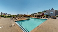 Located at 3200 Chestnut Ridge Drive in Pittsburgh, PA, this thoughtfully designed outdoor amenity area serves as a central gathering space within the community. The centerpiece is a generously sized swimming pool with clear blue water, framed by a broad concrete sundeck lined with numerous lounge chairs — ideal for relaxing under open skies. A elegant white pergola provides shaded seating near the pool, offering a comfortable retreat on sunny days.

Adjacent to the pool area, a raised terrace features picnic tables with yellow umbrellas, providing a dedicated outdoor dining and social space. The grounds are beautifully landscaped with a rich variety of ornamental grasses, flowering shrubs, manicured hedgerows, and colorful seasonal plantings that add vibrancy throughout. Winding pathways connect the various zones of the outdoor space, bordered by dense greenery that enhances privacy and creates a park-like atmosphere.

The surrounding residential buildings feature a classic combination of red brick and neutral siding exteriors with pitched rooflines, providing an attractive backdrop. Wooded hillsides visible beyond the property boundaries contribute to a natural, serene setting. The entire outdoor space benefits from abundant natural light, creating a bright and welcoming environment throughout the day.