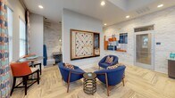 Located at 6255 Fernwood Dr. in Kannapolis, NC, this single-floor community clubhouse is thoughtfully designed to serve as a dynamic social hub. The space is anchored by a dramatic great room boasting soaring double-height ceilings painted a rich navy blue, illuminated by a striking contemporary chandelier. Oversized windows dressed in vibrant patterned curtains flood the interior with abundant natural light while offering views of the surrounding community.

A centerpiece billiards table with a bold blue felt surface commands the main lounge area, complemented by plush navy blue lounge chairs with colorful patterned accent pillows and bistro-style high-top tables with orange upholstered seating. The color palette of navy, orange, and teal is carried consistently throughout the space, creating a cohesive and energetic atmosphere.

A textured white stacked-stone accent wall serves as a backdrop for a wall-mounted flat-screen television above a sleek walnut media cabinet, providing a media lounge area ideal for group gatherings. An adjacent seating nook features a large framed Scrabble board wall display alongside contemporary artwork and additional blue lounge chairs arranged around a sculptural side table. Light wood-toned herringbone-pattern flooring ties the entire space together, lending warmth and a modern aesthetic throughout.