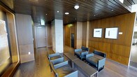This single-floor space exudes a refined, corporate aesthetic defined by its consistent use of rich wood paneling that lines both the walls and ceiling, lending warmth and a sense of understated luxury throughout. Recessed lighting and flush-mounted ceiling fixtures illuminate the interior evenly, highlighting the quality of the materials and the thoughtful design.

The main area functions as a waiting or reception lounge, furnished with a set of sleek, modern armchairs and side tables in dark metal and leather upholstery. The seating arrangement is orderly and deliberate, encouraging a composed and professional atmosphere. Hardwood flooring in a matching warm tone ties the space together cohesively.

A structural column clad in light stone or concrete provides a subtle contrast to the surrounding wood tones, adding an architectural focal point. Framed artwork on the paneled walls contributes a curated, gallery-like quality. Several doors are visible leading to additional rooms or corridors, suggesting the space is part of a larger facility. Overall, the design balances functionality with elegant, contemporary finishes to create a welcoming yet formal environment.