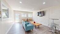 Located at 1039 North Alamo Avenue in Tucson, Arizona, this single-floor space offers a clean, modern design with a bright and functional layout. The living room serves as the heart of the space, finished with light gray wood-look flooring and crisp white walls that enhance the sense of openness. Recessed lighting throughout provides even, well-distributed illumination, while large windows along one wall invite generous natural light and offer views of an outdoor area with a pool visible beyond the glass entry door.

A standout feature of the living room is a textured three-dimensional accent wall finished in white, which adds architectural depth and visual interest. A wall-mounted flat-screen television is positioned on this feature wall, complementing the mid-century modern furnishings including tufted teal sofas, leather sling chairs, and a solid wood coffee table.

A compact kitchenette is neatly integrated into the space, outfitted with white shaker-style cabinetry, white subway tile backsplash, a stainless steel sink, and a round bistro-style table with bar stools — ideal for casual dining or a quick workspace.

Doors off the main living area lead to additional rooms, including what appears to be a dedicated fitness room equipped with exercise equipment, as well as a mirrored door that opens to a bathroom, adding to the overall utility of this well-appointed space.