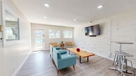 Located at 1039 North Alamo Avenue in Tucson, Arizona, this single-floor space offers a clean, modern design with a bright and functional layout. The living room serves as the heart of the space, finished with light gray wood-look flooring and crisp white walls that enhance the sense of openness. Recessed lighting throughout provides even, well-distributed illumination, while large windows along one wall invite generous natural light and offer views of an outdoor area with a pool visible beyond the glass entry door.

A standout feature of the living room is a textured three-dimensional accent wall finished in white, which adds architectural depth and visual interest. A wall-mounted flat-screen television is positioned on this feature wall, complementing the mid-century modern furnishings including tufted teal sofas, leather sling chairs, and a solid wood coffee table.

A compact kitchenette is neatly integrated into the space, outfitted with white shaker-style cabinetry, white subway tile backsplash, a stainless steel sink, and a round bistro-style table with bar stools — ideal for casual dining or a quick workspace.

Doors off the main living area lead to additional rooms, including what appears to be a dedicated fitness room equipped with exercise equipment, as well as a mirrored door that opens to a bathroom, adding to the overall utility of this well-appointed space.