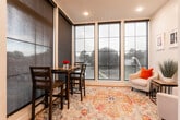 Located at 125 Bloomfield Avenue in the heart of Montclair, NJ, this single-floor space offers a thoughtfully designed and inviting environment that balances modern style with functional comfort. The most striking architectural feature is the expansive floor-to-ceiling window wall, which floods the interior with abundant natural light and provides open views of the surrounding streetscape and neighborhood, creating a lively connection to the outside world.

Warm-toned hardwood flooring runs throughout the space, adding a sense of richness and continuity. A cozy seating area anchors one corner of the room, complete with a plush white accent chair accented by a bold red pillow, a wood-and-metal side table, and a vibrant potted grass plant beneath a curated gallery of framed artwork. The arrangement is grounded by a colorful, patterned area rug that adds texture and warmth to the space.

Adjacent to the window wall, a high-top bistro table with two dark wood chairs serves as an intimate dining or meeting nook, perfectly positioned to take in the street views. Recessed lighting overhead complements the natural light, while a wall-mounted mini-split system ensures year-round comfort. The entry area is clean and functional, featuring a white door with a glass panel insert, an interior plant, and ample open floor space. Overall, the space exudes a bright, modern, and welcoming ambiance ideal for a variety of uses.