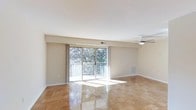This single-floor living space offers a clean, open layout filled with natural light. The main living area is anchored by polished parquet wood flooring laid in a classic block pattern, adding warmth and visual interest underfoot. Smooth white ceilings are equipped with a ceiling fan, promoting comfortable air circulation throughout the space. Large sliding glass doors dominate one wall, functioning as both a source of abundant natural light and a direct connection to an outdoor balcony. The balcony overlooks mature green trees, providing a pleasant, nature-filled backdrop. Neutral beige walls throughout create a versatile canvas well-suited to a variety of décor styles. A recessed beam detail near the sliding doors adds a subtle architectural element to the otherwise streamlined interior. Baseboard trim runs consistently along the walls, contributing to a tidy, finished appearance. The overall ambiance is bright, airy, and understated, making this an adaptable space ideal for comfortable everyday living.
