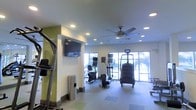 This fitness center is designed to provide a functional and comfortable workout environment. The space is finished with smooth, light-colored walls accented by a bold green feature wall, giving the room a clean and energetic feel. Recessed lighting throughout the ceiling, complemented by a ceiling fan, ensures the space remains well-lit and well-ventilated during workouts.

A range of cardio equipment is thoughtfully arranged across the floor, including a stair climber, elliptical machines, a stationary bicycle, and a stairmill-style machine, catering to a variety of fitness preferences and training goals. A wall-mounted flat-screen television is positioned prominently for entertainment during exercise sessions.

Large sliding glass doors and generously sized windows line one side of the space, flooding the interior with natural light and offering pleasant views of the surrounding greenery outdoors. The light-toned tile flooring is both durable and easy to maintain, practical for a high-use fitness environment. A sleek stainless steel waste bin adds a polished touch to the overall aesthetic.

The single-level layout ensures easy accessibility, making this fitness center a practical and inviting amenity for all users.