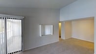 Located at 6200 Rieser Dr in Raleigh, NC, this single-story unit offers a straightforward, functional layout with clean, neutral finishes throughout. The main living area is highlighted by soaring vaulted ceilings that lend a sense of openness and airiness to the space, making it feel larger than its footprint suggests. The walls are painted in a soft, neutral gray tone that pairs well with the beige wall-to-wall carpeting underfoot, creating a cohesive and understated aesthetic. A built-in pass-through window is integrated into one wall of the living area, connecting it to an adjacent room and adding a practical architectural detail that enhances the flow between spaces. A sliding door — likely leading to a closet or exterior access — is visible along one wall, contributing to the room's storage or connectivity options. An open doorway leads further into the unit, where warm lighting hints at additional rooms beyond. The overall ambiance is calm and neutral, providing a blank canvas well-suited for personalization. The high ceilings remain a standout feature, giving the main living space an elevated, airy character uncommon in single-story layouts.