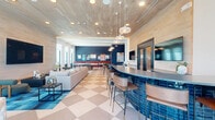 Located at 550 Westlake Park Blvd in Houston, TX, this contemporary amenity space spans a single floor and offers a thoughtfully designed environment ideal for socializing and recreation. The space is defined by a striking checkerboard-patterned tile floor that flows seamlessly throughout, tying together its distinct functional zones.

The lounge area features a generous sectional sofa, a large wall-mounted television, and a wood-paneled ceiling with recessed lighting that creates a warm, inviting ambiance. Adjacent to the lounge, a well-appointed bar area showcases vibrant blue tile accents and leather-topped bar stools, adding a bold pop of color and texture.

A dedicated game zone houses a full-size ping pong table set against floor-to-ceiling windows that flood the space with natural light and panoramic city views. Nearby, a dining nook with a round wooden table and sculptural upholstered chairs is illuminated by a statement chandelier, while floor-to-ceiling glass walls open the view to an outdoor pool terrace.

The hallway corridor, accented by a dramatic teal glazed tile feature wall and terracotta-hued lounge chairs, connects the various zones and leads to the outdoor terrace. The overall design palette — combining navy blue, warm wood, vibrant red accents, and neutral tones — achieves a modern, energetic aesthetic throughout.