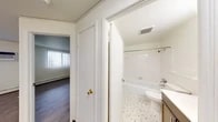 This single-floor unit offers a practical and well-organized layout that connects living, kitchen, and bathroom spaces in a straightforward flow. The main living area is generously proportioned, featuring wood-style flooring and large windows fitted with vertical blinds that allow natural light to fill the room. A baseboard heating system runs along the perimeter, providing consistent warmth throughout.

The kitchen is efficiently designed with dark brown cabinetry offering ample upper and lower storage, light countertops, a double-basin sink, and white appliances including a refrigerator and an electric range with overhead ventilation. The kitchen opens toward the main living space, creating an accessible and connected feel. A centrally located electrical panel is visible along one wall, and light wood-style flooring ties the area together.

The bathroom is finished in a clean, bright style with full white square tile surrounding the tub-shower combination. A mosaic tile floor with subtle green accents adds a classic touch. The space also includes a toilet, a vanity with storage, and a wall-mounted mirror. The unit's corridor connects the bathroom to the remaining rooms, maintaining a cohesive and functional layout throughout.