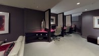 Located at 733 15th St NW in Washington, DC, this refined single-floor commercial suite offers a polished and professional environment designed with both comfort and function in mind. The space is defined by a rich, dark color palette of deep charcoal and taupe tones, accented throughout by bold magenta and crimson details that lend a distinctive, upscale character.

The lounge area anchors one end of the suite, featuring plush cream-upholstered seating, a sleek glass coffee table, and carefully curated framed artwork adorning the paneled walls. Recessed lighting and a table lamp provide a warm, inviting ambiance suited for client reception or informal meetings.

Transitioning deeper into the suite, a row of semi-private individual workstations is defined by elegant dark partitions with backlit panels. Each station is equipped with a computer and ergonomic task chair, set against vibrant magenta desk surfaces that contrast sharply with the dark cabinetry — a striking design choice that energizes the workspace.

A dedicated service alcove features built-in dark cabinetry with a magenta countertop housing a multifunction printer, complemented by a glass display shelf showcasing decorative vases. An additional seating nook with a black-and-white bench provides a transitional space near the suite entrance. Throughout, light carpet flooring, recessed ceiling lights, and paneled wall detailing maintain a cohesive, sophisticated aesthetic.