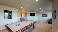 This single-floor game room is designed as a comfortable recreational space, combining entertainment amenities with a clean, modern aesthetic. The open layout is anchored by a full-size pool table with carved legs, positioned centrally on patterned low-pile flooring that adds texture while remaining practical for gameplay. A cue rack mounted to the wall ensures equipment stays organized and accessible.

The lounge area features dark leather armchairs arranged to encourage conversation or viewing, positioned near large windows dressed with horizontal blinds that allow natural light to filter into the space. Recessed ceiling lights supplement the natural light, providing consistent illumination throughout. A wall-mounted flat-screen television serves as a media focal point, ideal for casual viewing during gatherings.

A built-in wet bar area offers a stainless steel sink, a stone-patterned countertop, and contemporary wood-toned cabinetry above and below, making it convenient for hosting. A wall-mounted mini-split HVAC unit ensures year-round climate control. Decorative framed artwork and retro-style signage add personality to the neutral grey walls, completing the space with a relaxed, social atmosphere suited for recreation and entertainment.