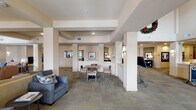 This single-floor communal facility offers a thoughtfully designed, open-plan interior that balances comfort and functionality across several distinct seating and activity areas. The space is anchored by a generously sized central lounge featuring plush sofas and armchairs in warm neutral tones, arranged around a coffee table to encourage conversation and relaxation. A highlight of the lounge is a fireplace flanked by framed artwork, creating a welcoming focal point. Structural columns with coffered ceiling details define zones within the open layout without fully enclosing them, lending the space an airy, flowing quality. A dedicated sitting nook near large, light-filled windows provides an intimate retreat with wingback chairs and a round side table, bathed in natural light filtered through horizontal blinds. Toward one section of the facility, a casual dining area features a round table with upholstered chairs, suitable for small group gatherings. Throughout, the flooring consists of patterned carpet tiles in muted, earthy tones that complement the warm beige and taupe wall palette. Wall sconces and recessed lighting provide a balanced, comfortable illumination scheme. The overall ambiance is calm, welcoming, and professionally appointed, consistent with a high-quality senior living or hospitality community common area.