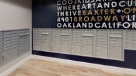 The mail area at 4901 Broadway in Oakland is a thoughtfully designed communal space that balances functionality with distinctive visual character. The room is finished with light wood-look plank flooring throughout, lending a clean and modern aesthetic underfoot. Neutral gray walls provide a calm backdrop, complemented by crisp white baseboards that frame the space neatly.

The most striking design element is a bold navy accent wall adorned with large typographic lettering in white and gold, referencing the building's address and community identity with phrases like "Where Art and Kids Thrive" and "4901 Broadway Oakland CA." This graphic wall treatment gives the space a strong sense of place and brand.

Multiple banks of recessed aluminum mailboxes line the walls, providing extensive mail and parcel storage capacity for residents. The mailboxes are organized across several wall-mounted units, including larger parcel compartments alongside standard letter slots. A dedicated recycling station with a wood-topped dark cabinet is conveniently positioned nearby. The ceiling features a coffered detail with recessed lighting, adding an architectural refinement that elevates the overall finish of this practical yet well-appointed space.