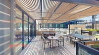 Located at 800 Van Cliburn Way in Fort Worth, Texas, this outdoor amenity space offers a thoughtfully designed courtyard environment centered around a resort-style swimming pool. The pool area is framed by clean-lined hardscape with wide concrete deck surrounds, lounge seating, and open sky views, all nestled within a modern multi-story building complex featuring brick and glass facades with private balconies above.

The courtyard is organized into distinct zones for relaxation and socializing. A generous turf lawn provides an open recreation area, bordered by raised planter beds filled with manicured shrubs and ornamental trees. Cozy seating clusters with plush lounge chairs are scattered throughout, accompanied by string lights that add ambiance for evening gatherings.

A standout feature is the covered outdoor dining and lounge pavilion, topped with a slatted wood pergola structure that filters natural sunlight into striking striped patterns across the large-format tile flooring below. Floor-to-ceiling glass walls on one side connect this covered zone to the interior, while open views to the courtyard maintain a seamless indoor-outdoor flow. Multiple dining tables and chairs furnish the space for group gatherings.

Additional pergola canopies with fabric shading provide relief from the Texas sun across the pool deck, completing a well-appointed, multi-functional outdoor retreat.