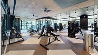 Located at 550 Westlake Park Blvd in Houston, Texas, this fitness center — branded as "The Watt" — offers a generously sized, single-floor exercise facility designed with both function and aesthetics in mind. The space is organized into distinct workout zones, separated by steel-framed glass partition walls that maintain an open, airy feel while defining dedicated areas for different training activities.

The flooring throughout features a bold geometric checkerboard pattern in neutral tones, providing visual interest while offering durable, cushioned surfaces suited for high-intensity use. Wood-paneled ceilings with oversized ceiling fans contribute warmth and ensure comfortable air circulation across the entire floor.

Floor-to-ceiling windows wrap around much of the perimeter, flooding the space with natural light and offering views of the surrounding greenery — creating an inviting atmosphere that sets this facility apart from typical gym environments. Pendant track lighting and recessed fixtures supplement the natural light in interior zones.

The equipment selection is comprehensive: multiple treadmills, elliptical machines, rowing machines, cable and resistance machines, free weights with a full dumbbell rack, adjustable benches, and a dedicated functional training area with turf flooring. Wall-mounted televisions and mirrors are strategically positioned throughout. Built-in shelving and a water station provide added convenience, making this a fully equipped fitness destination.