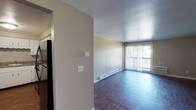 This single-floor unit is thoughtfully laid out to maximize functionality across its key living spaces. The living area serves as the central hub of the unit, featuring rich dark wood-style flooring, neutral-toned walls, and a sliding glass door fitted with vertical blinds that opens to a private balcony, allowing natural light to filter in. Baseboard heating runs along the perimeter, and a wall-mounted air conditioning unit provides additional climate control. The entry area connects seamlessly to the main living space via a straightforward corridor with a clean white front door.

The kitchen is separated from the living area by a partial wall, creating a defined yet accessible cooking space. It features white cabinetry, dark granite-style countertops, and black appliances including a refrigerator, electric range, and dishwasher, all set over matching dark wood-style flooring.

The bedroom offers a quieter retreat with soft wall-to-wall carpeting, neutral gray walls, a closet, and baseboard heating. The full bathroom is finished with wood-style flooring, a white vanity, and a tub-shower combination with brushed nickel fixtures and a curved curtain rod, giving it a clean, functional appearance throughout.