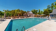 Located at 24 Winnow Walk Way in Wilmington, North Carolina, this outdoor community pool facility offers a well-appointed recreational retreat designed for relaxation and leisure. The centerpiece is a generously sized rectangular swimming pool with crystal-clear blue water, featuring clearly marked depth indicators and stainless steel entry rails for safe and easy access. The expansive concrete pool deck surrounds the pool on all sides, providing ample space for lounging and socializing. A variety of seating options are available throughout the deck, including cushioned lounge chairs, outdoor sofas, and shade umbrellas, creating comfortable gathering areas for residents. A dedicated poolhouse anchors one end of the facility, constructed with board-and-batten siding beneath a pitched shingle roof. The poolhouse provides essential amenities including restroom access, a water fountain, and an outdoor shower station for rinsing before and after swimming. Safety equipment, including a fire extinguisher, is prominently placed near the entrance. The entire pool area is enclosed by a black iron fence, ensuring security while maintaining open views of the surrounding mature trees and palm trees that lend a tropical ambiance. Under the expansive North Carolina blue sky, this facility presents a bright, inviting atmosphere ideal for outdoor recreation throughout the warmer months.