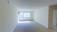 This single-floor space offers a clean, minimalist layout designed to maximize comfort and functionality. The living area is the centerpiece of the space, characterized by its bright, airy atmosphere thanks to a large window that allows generous amounts of natural light to flood the room. The window is fitted with vertical blinds, offering flexible light control and privacy. Soft, neutral-toned wall-to-wall carpeting lines the floor, adding warmth and a sense of calm to the space. The walls are finished in crisp white throughout, contributing to the open, uncluttered feel of the interior. A baseboard heating unit runs along the exterior wall beneath the window, ensuring efficient climate control during cooler months. A wall-mounted air conditioning unit provides additional comfort during warmer seasons. The overall ambiance is refreshingly simple and move-in ready, offering a blank canvas that can be easily personalized to suit a variety of design preferences. The space is well-suited for those who appreciate clean lines, abundant natural light, and a straightforward, functional living environment.