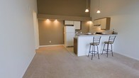 This single-floor space makes a strong first impression with its notably high ceilings, which lend an airy, expansive feel to the interior. The open-concept layout seamlessly connects the living area to the kitchen, creating a cohesive and functional floor plan well-suited for both relaxing and entertaining.

The living area is finished with wall-to-wall carpeting in a neutral beige tone, contributing to a warm and understated atmosphere. Warm taupe-colored walls run throughout, providing a consistent and grounded aesthetic across the space.

The kitchen is positioned at the rear of the open layout and features dark wood cabinetry paired with light upper cabinets, under-cabinet range hood ventilation, and a full-size refrigerator. A peninsula with a white countertop extends into the living space, accompanied by two metal bar stools, forming a convenient breakfast bar for casual dining.

Overhead, pendant lighting hangs from the ceiling above the kitchen area, supplemented by recessed lighting along the wall near the living space. The combination of high ceilings, open layout, and consistent neutral finishes creates a cohesive and inviting interior environment.