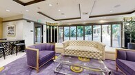 Located at 1026 S Broadway in the heart of Los Angeles, this striking single-floor living space exudes Hollywood glamour through its bold Art Deco-inspired design. The living room serves as the centerpiece, anchored by a distinctive purple, white, and gold circular-patterned carpet that sets a vibrant, theatrical tone throughout. Ornate black fluted columns with gilded Corinthian capitals frame the space, adding architectural drama and a sense of grandeur.

A white marble fireplace flanked by floor-to-ceiling mirrored panels and a wall-mounted flat-screen television creates a sophisticated focal point, while recessed lighting and a coffered ceiling contribute to the polished ambiance. Seating is both stylish and inviting, featuring a tufted cream sofa, plush purple velvet accent chairs, and a clear acrylic coffee table that adds a modern touch.

A grand black and gold piano occupies one corner, nodding to a creative, entertainment-forward lifestyle. A gallery wall of framed black-and-white celebrity portraits adds cultural character and a cinematic flair. Large sliding glass doors along one wall flood the space with natural light and offer views of lush outdoor greenery. A bar-height counter with mosaic tile detailing and dark stools provides a functional entertaining area near the entrance.