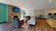 Located at 6701 W Carnation St in Richmond, VA, this single-story community amenity center offers a thoughtfully designed layout that balances comfort, functionality, and modern style. The space is accessed through a striking glass-panel entrance that opens into a bright, open interior with rich hardwood-style flooring throughout.

The lounge area serves as the social heart of the space, featuring a full brick fireplace wall with a wall-mounted TV, vibrant teal accent walls, and large glass doors that invite natural light and outdoor views. Comfortable contemporary seating, including a plush sofa and mid-century modern chairs, is anchored by a colorful patterned area rug.

A compact kitchenette provides everyday convenience, outfitted with white cabinetry, a sink, microwave, stainless steel refrigerator, and a wall-mounted TV — ideal for casual gatherings. Adjacent to this is a dining nook with natural wood communal tables, chartreuse upholstered chairs, pendant lighting, and a curated gallery of framed abstract artwork.

The fitness center is a standout feature, generously proportioned and well-equipped with treadmills, ellipticals, a stationary bike, free weights, stability balls, weight benches, and a large wall mirror. Teal accent walls, colorful athletic artwork, and large windows contribute to an energizing atmosphere. Light herringbone-pattern flooring and recessed lighting complete the clean, modern aesthetic throughout.