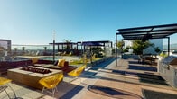 This expansive rooftop amenity deck is designed for elevated outdoor living, combining contemporary architecture with a resort-inspired atmosphere. The space is anchored by a large, sparkling swimming pool surrounded by yellow lounge chairs and synthetic turf strips, creating a vibrant, recreational setting under open skies. Multiple sleek pergolas provide shaded gathering areas throughout the deck, complementing the clean lines of the overall design. A dedicated lounge zone features plush sectional seating arranged around a central fire feature, with bold yellow accent chairs adding a striking pop of color. An outdoor kitchen and BBQ station with built-in grills offers a fully equipped space for entertaining. Warm wood-panel cladding on structural elements and privacy screens adds texture and visual warmth against the otherwise modern palette of concrete pavers and glass railings. Curated planters showcase drought-tolerant succulents and palms, bringing natural greenery to the rooftop environment. Overhead string lights in a dedicated garden corridor create an inviting evening ambiance. Access points clad in rich horizontal wood siding maintain a cohesive aesthetic throughout. Panoramic open-sky views and abundant natural sunlight define the overall character of this well-appointed rooftop retreat.