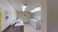 This shared laundry facility is designed with practicality and efficiency in mind, offering a clean and well-maintained environment for users. The room features two rows of white top-loading washing machines and dryers arranged in parallel lines along each wall, providing ample capacity for simultaneous use. Each unit is neatly spaced and easily accessible, streamlining the laundry process. The space is finished with neutral white walls and light-toned ceramic floor tiles, contributing to a bright and hygienic atmosphere. Overhead fluorescent lighting fixtures are evenly distributed across the ceiling, ensuring the entire room is well-lit. A posted notice board on the wall provides laundry room rules and relevant information for users. The layout is open and uncluttered, allowing for easy movement between machines. The single-floor facility is straightforward in its design, prioritizing function and convenience while maintaining a tidy and orderly appearance throughout.