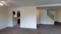 This two-floor residence offers a practical and well-organized layout designed for comfortable everyday living. The main level features an open-concept design that seamlessly connects the living area to the kitchen, creating a cohesive and functional space. The living area is finished with plush carpeting that extends throughout, adding warmth and comfort underfoot.

The kitchen is equipped with dark cabinetry that provides ample storage, a gas range with a white finish, and a sink positioned beneath a window that allows natural light to filter in. The contrast between the dark cabinets and light countertops and walls gives the kitchen a clean, defined aesthetic.

A partial dividing wall separates the living space from the entryway area, where a closet with louvered doors offers convenient storage. A staircase leads to the upper floor, rounding out the main level's layout. The walls throughout are finished in crisp white, contributing to a bright and neutral atmosphere that is easy to personalize. Overall, the space presents a straightforward and livable design with a clear separation of functional zones across both floors.