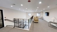 This upper-level space features a clean, open layout with neutral white walls and wall-to-wall carpeting that creates a warm, understated ambiance. A prominent staircase with sleek black metal railings serves as a central architectural element, providing both structural definition and visual interest to the room. The stairwell opening is bordered by a matching railing system that maintains an open, airy feel while clearly delineating the transition between levels.

The ceiling is outfitted with an array of recessed lighting fixtures, ensuring consistent, even illumination throughout the space. A fire sprinkler head is also visible at the ceiling's center, reflecting practical safety considerations. Two windows along the far wall allow natural light to filter into the room, complementing the artificial lighting and brightening the overall environment.

In the lounge area adjacent to the staircase, a beige sofa and matching armchair are arranged to create a comfortable seating space. A small side table with a lamp adds functional detail to this corner. The overall design is straightforward and functional, with a minimalist aesthetic that makes the space feel open and well-maintained.