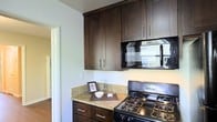 This single-story space includes a thoughtfully designed kitchen that serves as a functional and stylish centerpiece. The kitchen features rich, dark espresso-toned cabinetry with clean shaker-style doors and sleek bar-pull hardware, extending from floor to ceiling to maximize storage capacity. The countertops are finished in light granite, offering a striking contrast against the dark cabinetry while providing a durable and elegant work surface. A black gas range with multiple burners anchors the cooking area, complemented by an over-the-range microwave with a built-in ventilation hood. The cabinetry layout includes both upper and lower units, offering generous storage throughout. Light-toned walls keep the space feeling bright and open, while a doorway visible in the background hints at additional living areas beyond the kitchen. Natural light filters in through a nearby window, contributing to the overall welcoming atmosphere. The overall design aesthetic balances practicality with visual appeal, combining classic finishes with modern fixtures for a cohesive and inviting interior environment.