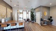 This well-appointed single-floor communal space masterfully combines modern design with warm, rustic elements to create an inviting and functional environment. The open layout flows seamlessly between several distinct areas, each thoughtfully designed to serve a specific purpose while maintaining a cohesive aesthetic throughout.

The main reception and gathering area features light hardwood-style flooring, recessed lighting, and a central wooden bar-height table with bar stools, anchored by a geometric-patterned area rug. A dramatic dark accent wall serves as a focal point, complemented by paired pendant lights and decorative wall art. Large-framed sliding barn-style doors with frosted glass panels add an industrial-chic touch while allowing flexible division of the space.

A comfortable lounge area offers a vibrant teal sofa, leather accent chairs, and a live-edge wood coffee table set atop a bold black-and-white patterned rug. A large abstract teal canvas painting adorns the wall, and a ceiling-mounted flat-screen television adds a modern amenity.

The dedicated business center features a built-in white countertop workstation with a computer and printer, set against a striking exposed brick wall with an arched transom window that floods the area with natural light. WiFi is available throughout.

Tall indoor plants, French doors opening to an outdoor patio, and the interplay of brick, wood, and contemporary finishes give this space a warm yet modern character.