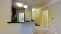 This single-floor residence showcases an open-concept layout that seamlessly connects the kitchen and living areas. The kitchen is anchored by a curved peninsula with a dark granite or stone countertop, providing both prep space and a casual bar-style seating area. Dark cabinetry contrasts with the light-colored walls, while a stainless steel refrigerator is visible in the background. Two elegant pendant lights hang above the peninsula, complementing the flush ceiling fixture to create layered, functional lighting throughout the space.

Crown molding runs along the ceiling, adding a refined architectural detail that elevates the overall aesthetic. The flooring transitions from light-toned carpet in the living area to a harder surface in the kitchen zone. Neutral, warm-toned walls contribute to a bright and welcoming ambiance. Multiple doorways are visible from this vantage point, suggesting a well-connected floor plan with easy access to adjacent rooms and a hallway. The layout appears efficient and thoughtfully designed, offering a comfortable flow between the cooking, dining, and living spaces within a compact yet functional footprint.