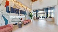 Located at 14300 Boggy Creek Rd in Orlando's sought-after Lake Nona community, this striking single-floor lobby space makes an immediate impression with its soaring double-height ceilings and expansive, light-filled interior. Large-format porcelain tile flooring runs throughout, reflecting the abundant natural light that pours in through floor-to-ceiling glass walls and doors.

A dramatic, large-scale artistic mural anchors the entry area, accompanied by a glowing neon "Lake Nona – Say You're Here" sign and a plush pink sofa, setting an energetic and welcoming tone. The open layout flows into a generous lounge area featuring a sculptural curved white sofa, a suspended modern fireplace framed by a bold geometric-patterned accent wall, and colorful art pieces that add personality throughout.

A sleek coffee and refreshment bar with warm wood cabinetry, open shelving, and a built-in beverage cooler provides a functional yet stylish amenity. Glass-panel sliding doors lead to an adjacent private meeting or conference room. Striking globe chandeliers and a slatted wood ceiling detail in the entry add warmth and elegance overhead.

Floor-to-ceiling windows frame views of a lush outdoor pool area with palm trees, blurring the boundary between interior comfort and Florida's outdoor lifestyle. The space's design palette — bold navy blue accents, crisp white walls, and natural wood tones — creates a cohesive, contemporary ambiance throughout.