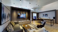 This co-working lounge is a thoughtfully designed, single-floor space that blends professional functionality with a welcoming, upscale aesthetic. The soaring ceilings immediately establish a sense of openness, enhanced by a row of clerestory windows that draw in generous amounts of natural light along the upper perimeter of the space.

The lounge is organized into distinct zones to accommodate a variety of working and social needs. A comfortable seating area anchors the main space, featuring a plush sofa and armchairs arranged around a wooden coffee table atop a textured area rug, ideal for casual meetings or relaxed individual work. A large wall-mounted flat-screen television serves as a focal point against a dark accent wall framed by recessed display cabinetry with clean, modern hardware.

Adjacent to the lounge area, a bar-height communal table with industrial-style stools offers an alternative workspace suited for quick tasks or informal collaboration. A separate room visible through an open passage provides additional seating, giving occupants the option of a quieter, more private environment. Pendant lighting and track fixtures throughout contribute to the polished, contemporary atmosphere of the overall space.