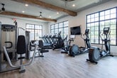 Located at 7243 US-431 N in Owens Cross Roads, Alabama, this single-story fitness center offers a thoughtfully designed workout environment that balances modern functionality with warm, rustic aesthetics. Exposed wood beams traverse the elevated ceilings throughout the space, complemented by ceiling fans and recessed lighting that create a bright, comfortable atmosphere.

Large, grid-style black-framed windows line multiple walls, flooding the interior with natural light while providing views of the surrounding outdoor area, including a pool and adjacent building. The light-toned hardwood-style flooring enhances the open, airy feel of the space.

The cardio zone is well-appointed with multiple treadmills, elliptical trainers, and stationary bikes, all positioned to take advantage of the expansive window views. The strength training area features Precor cable machines, a power rack, a full array of kettlebells and free weights, a punching bag, and adjustable benches. A large wall-mounted mirror extends the visual depth of the room, while a built-in shelving unit along one wall provides organized storage for medicine balls, accessories, and equipment. The overall ambiance is clean, spacious, and well-suited for a comprehensive fitness experience.