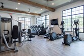 Located at 7243 US-431 N in Owens Cross Roads, Alabama, this single-story fitness center offers a thoughtfully designed workout environment that balances modern functionality with warm, rustic aesthetics. Exposed wood beams traverse the elevated ceilings throughout the space, complemented by ceiling fans and recessed lighting that create a bright, comfortable atmosphere.

Large, grid-style black-framed windows line multiple walls, flooding the interior with natural light while providing views of the surrounding outdoor area, including a pool and adjacent building. The light-toned hardwood-style flooring enhances the open, airy feel of the space.

The cardio zone is well-appointed with multiple treadmills, elliptical trainers, and stationary bikes, all positioned to take advantage of the expansive window views. The strength training area features Precor cable machines, a power rack, a full array of kettlebells and free weights, a punching bag, and adjustable benches. A large wall-mounted mirror extends the visual depth of the room, while a built-in shelving unit along one wall provides organized storage for medicine balls, accessories, and equipment. The overall ambiance is clean, spacious, and well-suited for a comprehensive fitness experience.