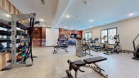 Located at 346 W 26th St in Charlotte, NC, this fitness center offers a generously sized, single-floor exercise space designed to accommodate a wide variety of workout routines. The room features polished concrete flooring throughout, providing a durable and easy-to-maintain surface suited for high-traffic use.

The space is organized into distinct functional zones. The cardio area, positioned along the windowed wall, houses multiple treadmills, elliptical machines, and a stair climber, benefiting from natural light filtering through several windows. A dedicated strength training zone features a multi-station cable machine, a Smith machine with bench press setup, and adjustable weight benches. A neatly organized dumbbell rack lines one wall alongside floor-to-ceiling mirrors, enhancing the sense of space and allowing users to monitor their form.

A striking, large-scale artistic mural in warm red and amber tones serves as a bold focal point on one wall, adding visual energy to the environment. Recessed lighting and a tray ceiling detail contribute to the polished, well-lit atmosphere. Additional amenities include a wall-mounted television, a water fountain station, and restroom access, making this a fully functional and thoughtfully appointed fitness facility.
