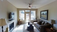 This single-floor space offers a warm and inviting atmosphere highlighted by thoughtful design and functional comfort. The living room serves as the centerpiece, bathed in natural light streaming through a set of large, multi-pane windows flanking a classic white paneled door that opens to the outdoors. A ceiling fan with a light fixture is centrally mounted, ensuring year-round comfort. The room features a tiled fireplace along one wall, adding a cozy focal point, while a wall-mounted flat-screen television completes the entertainment setup. Light-toned tile flooring extends throughout, complemented by a decorative area rug that anchors the seating arrangement. The living space is furnished with a plush, earth-toned sofa accented with patterned throw pillows, paired with a dark wood coffee table. A large, framed landscape painting adorns one wall, adding a refined artistic touch to the neutral beige color palette. Decorative elements such as a tall vase with branches and woven storage baskets contribute to the space's understated, classic aesthetic. Views through the windows reveal lush greenery outside, connecting the interior to the natural surroundings and enhancing the overall sense of openness and tranquility.