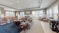 This well-appointed single-floor space offers an open and inviting layout designed for both comfort and functionality. The centerpiece is a generously sized living area featuring a tray ceiling with recessed lighting and elegant crown molding. A stone-surround fireplace flanked by built-in wood cabinetry and a mounted television serves as the room's focal point, while plush seating arranged over a rich navy area rug creates a warm, social atmosphere. Large windows dressed with white blinds flood the space with natural light and offer views of the surrounding neighborhood.

The dining area is seamlessly integrated into the open floor plan, featuring a dark wood table set against a wall of windows framed by classic wainscoting — a detail that carries throughout the space, lending a refined coastal character.

The kitchen is thoughtfully designed with warm natural wood cabinetry, marble-look countertops, a center island, and stainless steel appliances including a full-size refrigerator. Tile flooring, ample counter space, and a bright window over the sink make it both practical and pleasant. An adjacent utility area with additional cabinetry, a built-in oven, and a stainless sink provides extra workspace. The overall palette of soft grays, warm woods, and crisp white trim creates a cohesive and welcoming environment throughout.