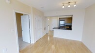 This single-floor residence offers a thoughtfully designed open-concept layout that seamlessly connects the living and kitchen areas. Light hardwood flooring runs throughout the main living space, lending warmth and a sense of continuity to the interior. The kitchen is positioned at the heart of the home, featuring dark granite-style countertops, medium-toned wood cabinetry, stainless steel appliances including a refrigerator, and a peninsula with a built-in sink — ideal for both meal preparation and casual entertaining. A track lighting fixture overhead provides focused illumination across the space. The soft, neutral wall tones in warm beige complement the light flooring and white trim, creating a clean and inviting ambiance. Multiple interior doors visible from the main area suggest well-defined private spaces, including what appears to be a closet and additional rooms beyond. The layout is efficient and well-organized, making excellent use of the available footprint. Overall, the space presents a bright, functional, and move-in-ready environment suitable for everyday comfortable living.