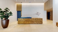 Located at 215 Grove Street in Jersey City, NJ, this is the lobby hallway of One Grove — a thoughtfully designed entrance space that sets an elegant tone for the building. The layout is open and generously proportioned, with large-format light-colored tile flooring that extends throughout, lending a clean and polished aesthetic.

At the heart of the lobby stands a striking reception desk crafted from natural reclaimed wood, featuring a rich grain texture and a deep blue countertop that provides a bold, modern contrast. Behind the desk, vertical wood slat panels frame a vivid blue door, adding depth and warmth to the otherwise crisp white walls. Recessed track lighting and a softly glowing LED cove light along the ceiling create a layered, ambient illumination that enhances the space's contemporary character.

The lobby is accented with curated greenery, including a sculptural bonsai tree and a tall fiddle-leaf fig in a dark rounded planter, introducing organic elements that complement the natural wood tones. A low rustic wood bench near the entrance offers a functional yet stylistic touch.

Floor-to-ceiling glass at the building entrance floods the space with natural light and provides clear views to Grove Street, while a dedicated mailbox area with a wood slat surround adds practical functionality. The overall ambiance is modern, refined, and welcoming.