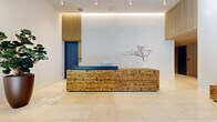 Located at 215 Grove Street in Jersey City, NJ, this is the lobby hallway of One Grove — a thoughtfully designed entrance space that sets an elegant tone for the building. The layout is open and generously proportioned, with large-format light-colored tile flooring that extends throughout, lending a clean and polished aesthetic.

At the heart of the lobby stands a striking reception desk crafted from natural reclaimed wood, featuring a rich grain texture and a deep blue countertop that provides a bold, modern contrast. Behind the desk, vertical wood slat panels frame a vivid blue door, adding depth and warmth to the otherwise crisp white walls. Recessed track lighting and a softly glowing LED cove light along the ceiling create a layered, ambient illumination that enhances the space's contemporary character.

The lobby is accented with curated greenery, including a sculptural bonsai tree and a tall fiddle-leaf fig in a dark rounded planter, introducing organic elements that complement the natural wood tones. A low rustic wood bench near the entrance offers a functional yet stylistic touch.

Floor-to-ceiling glass at the building entrance floods the space with natural light and provides clear views to Grove Street, while a dedicated mailbox area with a wood slat surround adds practical functionality. The overall ambiance is modern, refined, and welcoming.