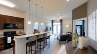 This single-floor residence showcases an open-concept layout that seamlessly connects the kitchen and living areas into one cohesive, well-lit space. The kitchen features a granite-topped breakfast bar with seating for multiple guests, served by three elegant pendant lights that hang at staggered heights, providing both functional illumination and a stylish focal point. Dark cabinetry adds a rich contrast to the light-toned walls and countertops.

The living area extends naturally from the kitchen and dining space, anchored by a ceiling fan that enhances air circulation throughout. French doors at the far end of the room invite generous natural light and offer a view to an outdoor area, contributing to the airy, open ambiance. Neutral-toned walls and light flooring create a cohesive backdrop throughout the space.

A dedicated living zone features a comfortable seating arrangement centered around a glass coffee table, with a television console positioned against the wall. The overall design balances practicality with modern aesthetics, making the space feel both inviting and functional for everyday living and entertaining.