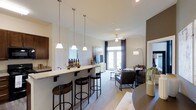 This single-floor residence showcases an open-concept layout that seamlessly connects the kitchen and living areas into one cohesive, well-lit space. The kitchen features a granite-topped breakfast bar with seating for multiple guests, served by three elegant pendant lights that hang at staggered heights, providing both functional illumination and a stylish focal point. Dark cabinetry adds a rich contrast to the light-toned walls and countertops.

The living area extends naturally from the kitchen and dining space, anchored by a ceiling fan that enhances air circulation throughout. French doors at the far end of the room invite generous natural light and offer a view to an outdoor area, contributing to the airy, open ambiance. Neutral-toned walls and light flooring create a cohesive backdrop throughout the space.

A dedicated living zone features a comfortable seating arrangement centered around a glass coffee table, with a television console positioned against the wall. The overall design balances practicality with modern aesthetics, making the space feel both inviting and functional for everyday living and entertaining.