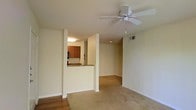 This single-floor residence offers an open and functional layout designed for comfortable everyday living. The main living area is finished with warm carpeting and neutral walls, creating a welcoming atmosphere throughout. A ceiling fan is centrally mounted overhead, providing both practical air circulation and a subtle design element to the space.

A defining architectural feature is the half-wall pass-through that connects the living area to the kitchen, allowing for an open sightline between the two spaces while maintaining a sense of definition. The kitchen is equipped with warm wood-toned cabinetry and granite-style countertops visible over the pass-through bar, alongside built-in appliances including a microwave. Recessed lighting in the kitchen provides focused illumination, complementing the natural light that filters throughout the unit.

The transition between the entryway and the main living space is marked by a shift from hardwood-style flooring to plush carpet, adding textural variety underfoot. HVAC venting is integrated into the ceiling, ensuring climate comfort year-round. The overall color palette — soft whites, warm beiges, and natural wood tones — contributes to a clean, neutral aesthetic that is both inviting and versatile.