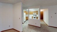 This single-floor residential unit offers a practical and well-organized layout designed for comfortable living. The living area is generously sized, featuring neutral-toned carpet, white walls with wood baseboards, a textured ceiling, and a wall-mounted air conditioning unit. A sliding glass door leads to a private balcony, allowing natural light to fill the room and providing outdoor views of surrounding trees.

The kitchen is positioned adjacent to the living area and is visible through a pass-through opening, creating an open and connected feel. It is equipped with warm oak cabinetry — both upper and lower — white appliances including a refrigerator, electric range, and dishwasher, along with white countertops and a double stainless steel sink set into a corner layout.

The bedroom includes plush carpeting, a ceiling fan with light fixture, wood-trimmed windows with outdoor views, and a baseboard heating unit. The room is well-proportioned and benefits from natural light.

The bathroom features tile flooring in a stone-like pattern, a full tub-shower combination with white tile surround, a toilet, and a vanity with a light countertop and wood cabinetry. The overall aesthetic throughout the unit is clean and neutral, offering a move-in-ready canvas with functional, classic finishes.