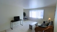This single-floor residence offers a clean, functional layout with an emphasis on open living. The living area is spacious and well-lit, anchored by plush carpeting and a large picture window dressed with vertical blinds that allow an abundance of natural light to fill the room. The minimalist design is complemented by smooth white walls and a neutral color palette, creating a calm and airy atmosphere throughout. A modest living setup accommodates a flat-screen television on a sleek console table, a slatted metal coffee table, and comfortable seating, making the space practical for everyday use. The living area flows seamlessly into a dedicated dining space, which features a compact dining table and chairs, partially separated by an open partition wall. A lush indoor plant adds a touch of greenery to the dining nook, softening the otherwise clean, contemporary aesthetic. The continuous carpeting and consistent finishes across both areas reinforce a cohesive and unified design. Overall, the space presents a straightforward, light-filled layout well-suited for comfortable everyday living.