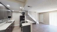 This two-story residence offers a thoughtfully designed layout with modern finishes throughout. The main level centers around an updated kitchen equipped with dark espresso cabinetry, white quartz-style countertops, a mosaic tile backsplash, stainless steel appliances including a range with overhead hood vent and refrigerator, and recessed lighting. A breakfast bar provides additional counter space and connects seamlessly to the open living area. The living room features durable wood-look flooring, neutral-toned walls, a textured ceiling, and floor-to-ceiling vertical blinds that allow generous natural light to fill the space. A staircase with white railings leads to the upper level, where the bedrooms are finished with plush carpeting, soft neutral walls, and distinctive exposed beam ceilings with white-painted wood paneling — a standout architectural detail that adds character and warmth. Windows with horizontal blinds bring in natural light and exterior views. The main-level bathroom offers a sleek walk-in shower with sliding glass doors and chrome fixtures, while a second bathroom on the upper level features a full soaking tub and shower combination. Wood-look flooring carries through the bathrooms, tying together the home's cohesive, contemporary aesthetic.